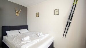 Uma cama bem arrumada com lençóis e toalhas brancos, uma cabeceira escura, uma cabeça de veado montada e duas imagens emolduradas na parede.