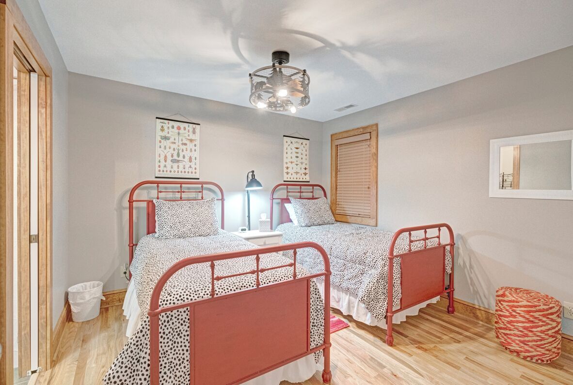Une chambre avec deux lits en métal, un luminaire au plafond, un plancher en bois et une fenêtre avec des stores.