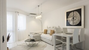 A modern living room with a white sofa, a glass coffee table, and a dining table with chairs. There is a chandelier hanging from the ceiling and a large abstract painting on the wall. - Scandic - City Center by HD Properties (Vilamoura)