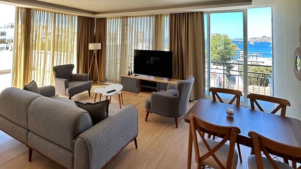 Un salon moderne comprenant un canapé, des fauteuils, une table à manger et un téléviseur. De grandes fenêtres offrent une vue sur la mer.
