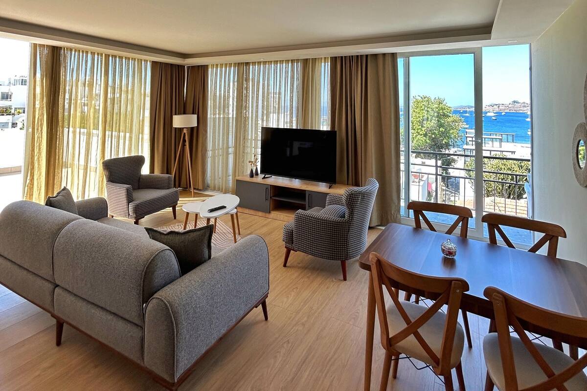 Un salon moderne comprenant un canapé, des fauteuils, une table à manger et un téléviseur. De grandes fenêtres offrent une vue sur la mer.