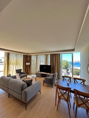 Un salon moderne comprenant un canapé, des fauteuils, une table à manger et un téléviseur. De grandes fenêtres offrent une vue sur la mer.