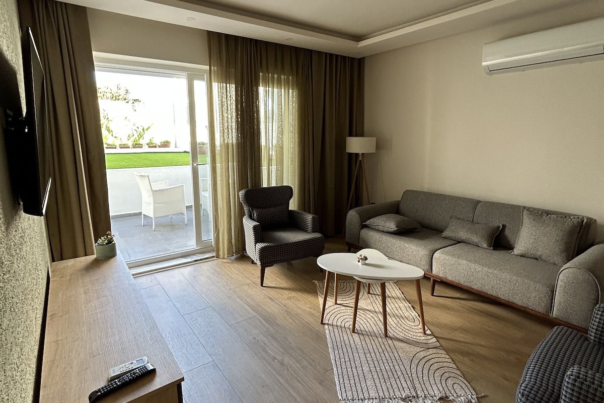 Un salon moderne avec un canapé, un fauteuil et une petite table basse. On y trouve un téléviseur posé sur un meuble, une télécommande sur une table en bois et une fenêtre avec des rideaux.