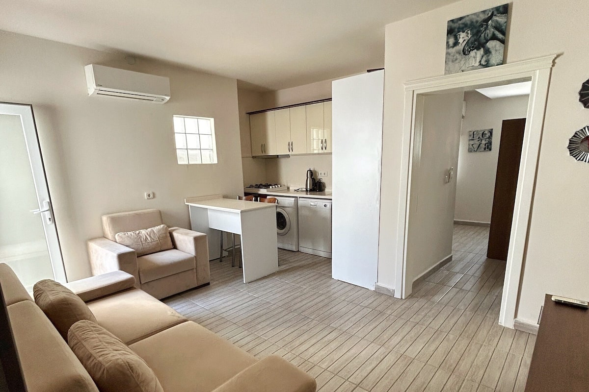 Un salon moderne avec un canapé beige, un coin repas doté d’une petite table et une cuisine avec des meubles blancs.