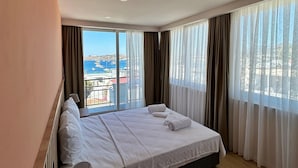 Une chambre d’hôtel dotée d’un grand lit, d’une table de chevet avec une lampe et offrant une vue sur la ville grâce à de grandes fenêtres du sol au plafond.
