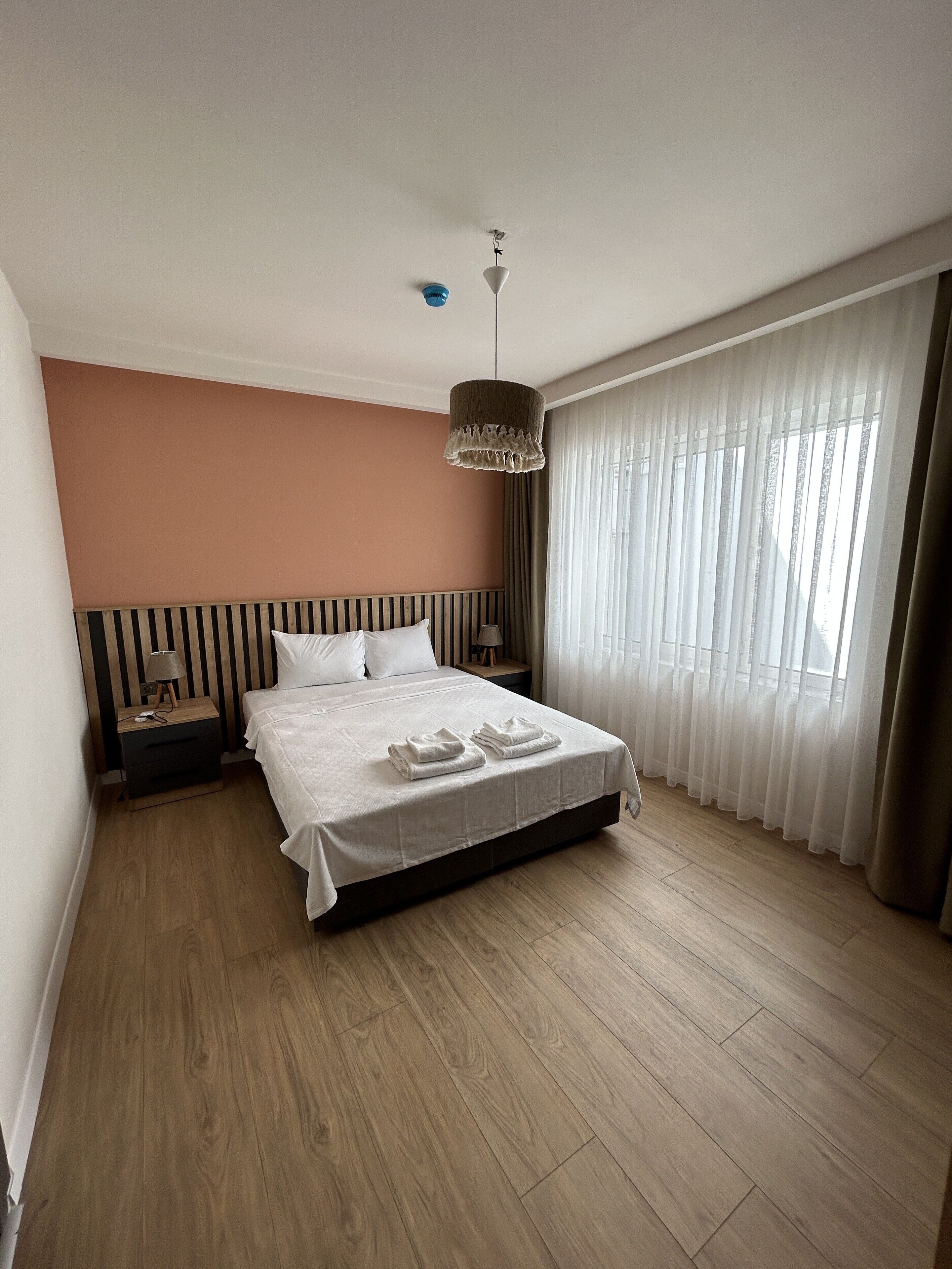 Una cama bien hecha con una colcha blanca y dos toallas dobladas, una mesita de noche con una lámpara, una mesita auxiliar de madera, una lámpara colgante y cortinas translúcidas en una ventana.