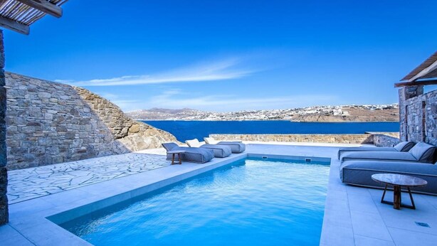 A pool area with stone walls, a view of the sea, and outdoor seating. - Mykonos Town Villa (Mykonos)