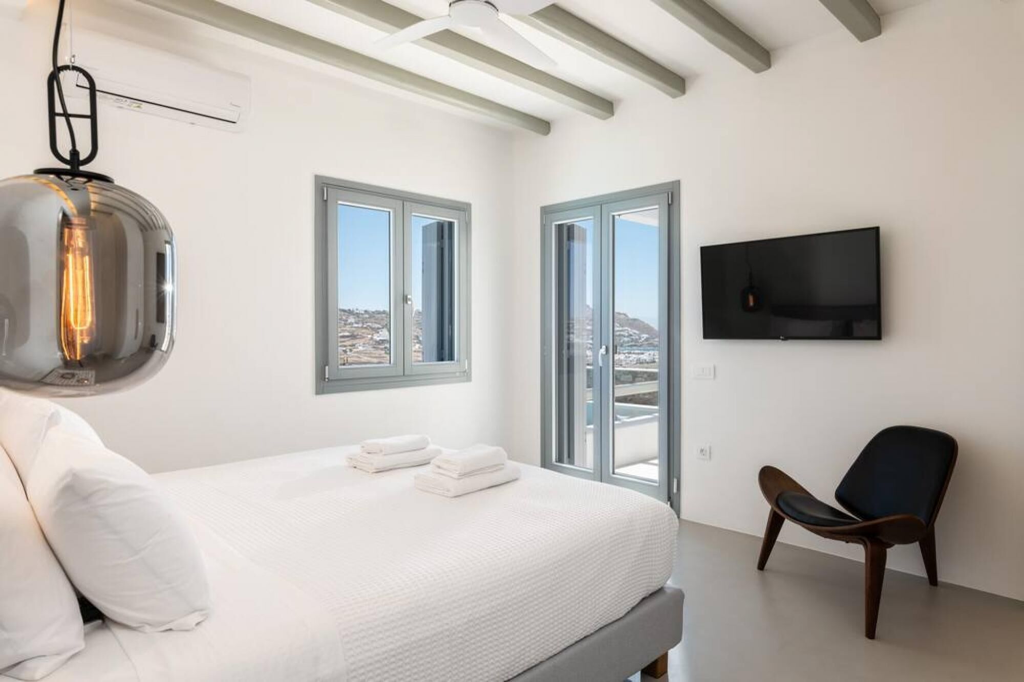 A bedroom with a bed, a chair, a TV, and large windows.