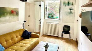 Un salon avec un canapé jaune, une table sur laquelle est posée une plante, un radiateur et une fenêtre donnant sur un jardin.