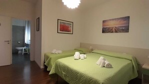A hotel room with two beds, each with green bedding and towels. There are two framed pictures on the walls, one depicting a landscape and the other a sunset. The room has a wooden floor and a door leading to a bathroom with a toilet and sink. - La Rosa dei Venti (Ortona)