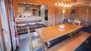 Ein gemütliches Holzinterieur mit Essbereich, der einen langen Tisch und Bänke umfasst, einer Küche mit modernen Geräten sowie einer Sitzecke mit Sofa und Sesseln.