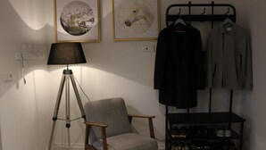 Een kamer met een driepotige vloerlamp, een grijze fauteuil, een kapstok met kleding en een schoenenrek met meerdere schoenparen.