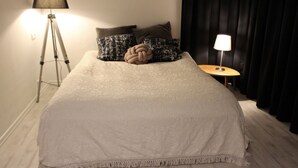 Een net opgemaakt bed met een witte deken en een decoratief knoopkussen, een vloerlamp en een nachtkastje met een lamp.