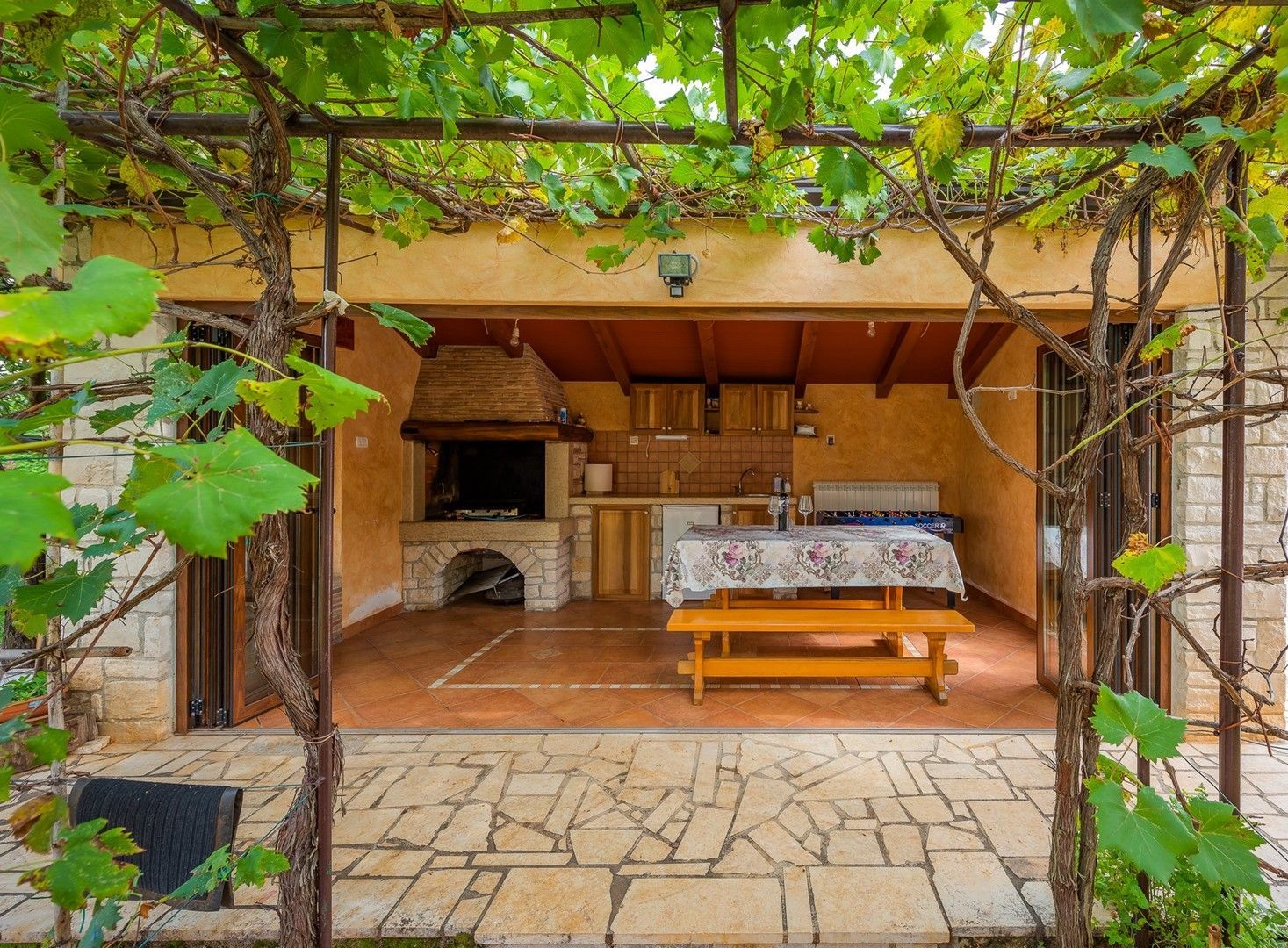 Una cocina rústica al aire libre con chimenea de piedra, mesa y banco de madera, rodeada por una pérgola con plantas trepadoras.