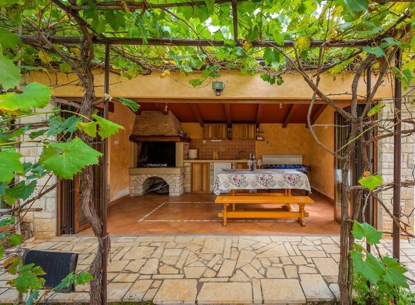 Una cocina rústica al aire libre con chimenea de piedra, mesa y banco de madera, rodeada por una pérgola con plantas trepadoras.