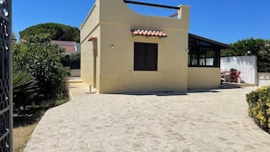 A modern building with a flat roof, a small attached structure with a tiled roof, and a paved entrance area. - Vacation home Andreina (the yellow) (Ognina)