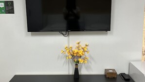 A hotel reception area with a flat-screen TV mounted on the wall, a wooden reception desk, a vase with yellow flowers, a pair of slippers, and a small wooden box. - IO hotel Bishkek (Bishkek)