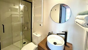 Un baño moderno con ducha envuelta en vidrio, inodoro, lavamanos de forma redonda, espejo y toallero.