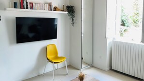 Un salón moderno con televisor de pantalla plana montado en la pared, una butaca amarilla, un mueble blanco y una ventana con vistas a la vegetación.