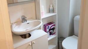 Ein Badezimmer mit weißem Waschbecken, Spiegel und Toilette. Ein Ablagebereich beinhaltet Pflegeprodukte und einen Haartrockner.
