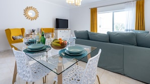Un soggiorno moderno con tavolo da pranzo in vetro, sedie bianche, un divano e una TV su un mobile bianco.