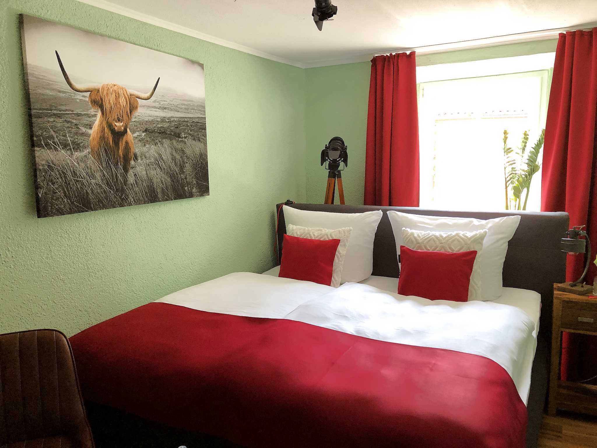 Une chambre à coucher avec un lit, une literie rouge et blanche, un tableau représentant une vache des Highlands accroché au mur, un trépied et une fenêtre avec des rideaux rouges.