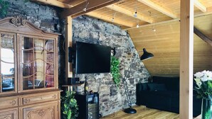 A rustic living room with a stone wall, wooden ceiling beams, a TV mounted on the wall, a cabinet with glass doors, a wine cooler, and a black sofa.