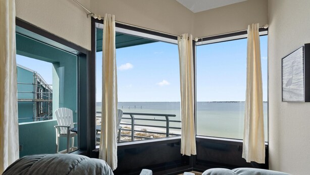 A room with a large window overlooking a balcony with a view of the sea. There is a chair and a sofa inside the room. - Escape to paradise at Palm Beach Club #217/Sound Front Condo! (Pensacola Beach)