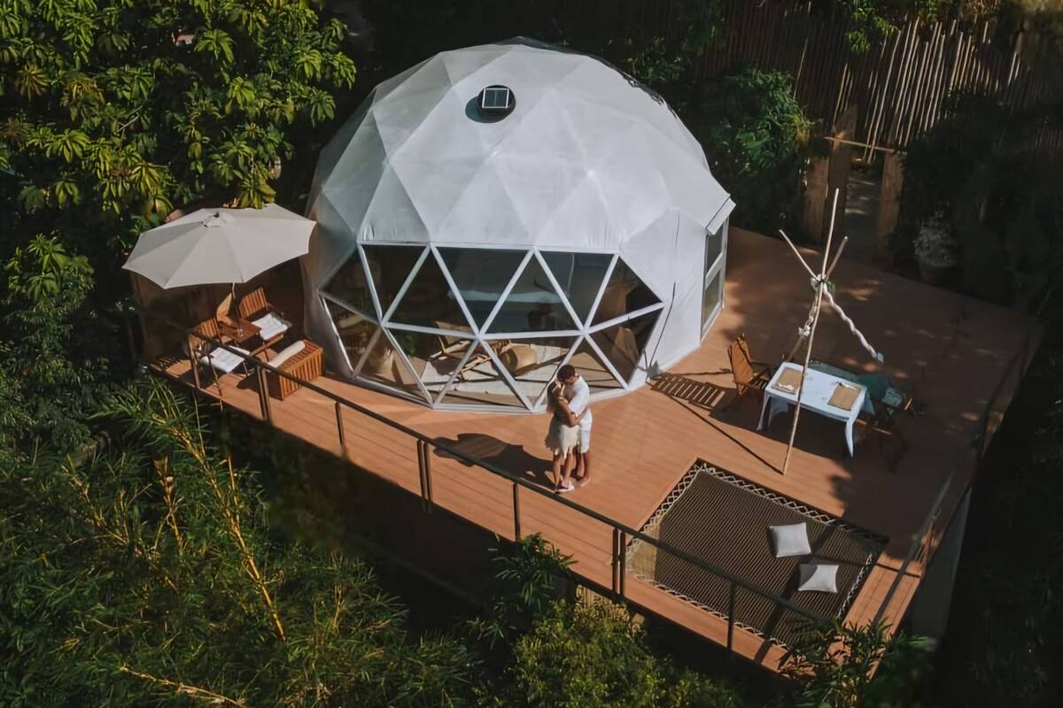 Eine geodätische Kuppel mit einem Oberlicht in der Mitte, umgeben von einer Holzterrasse mit Sitzgelegenheiten und einer Hängematte.