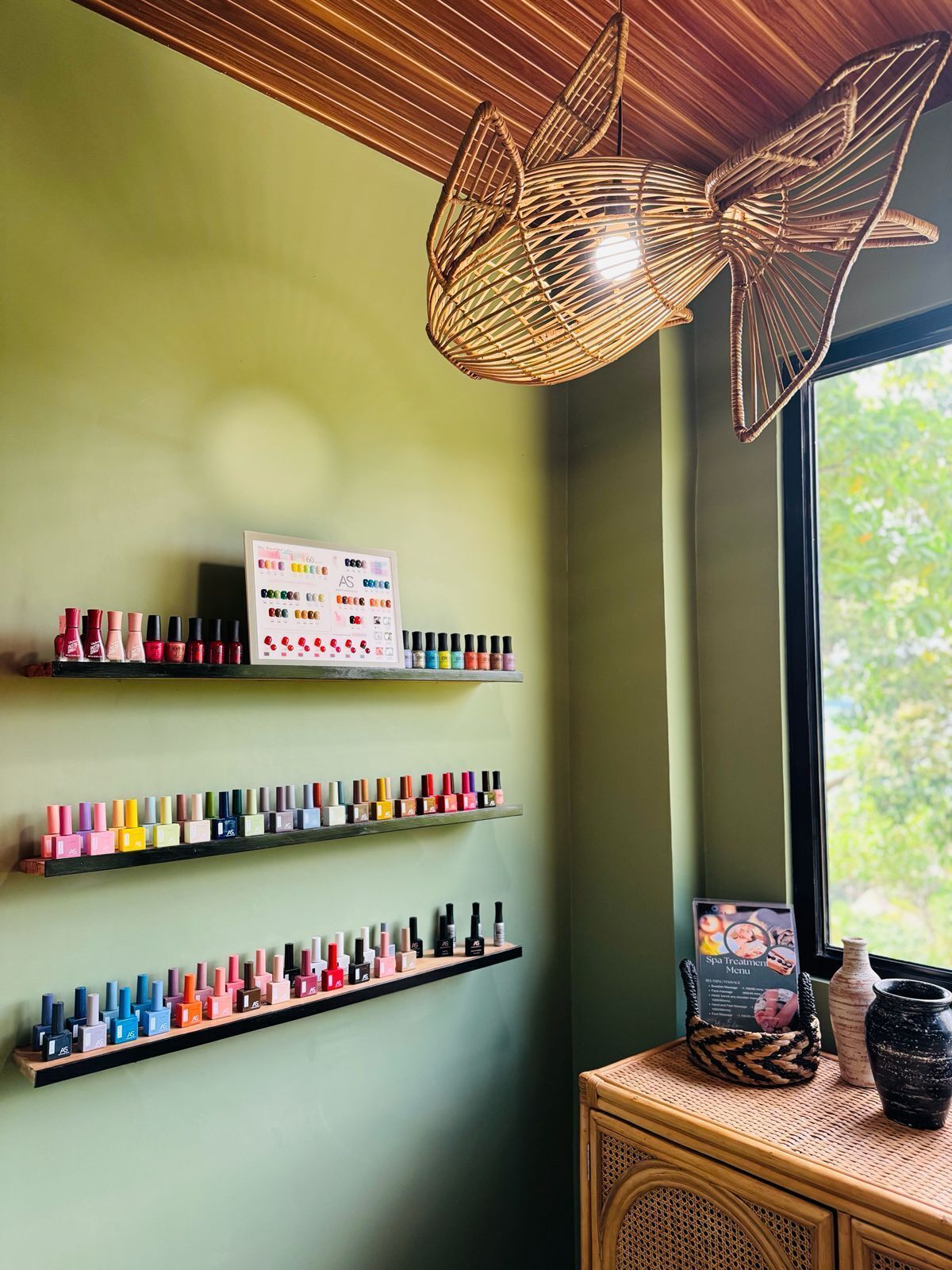 Una habitación con estantes que exhiben botellas de esmalte de uñas, un cartel con colores de esmalte, una lámpara de fibra natural y una ventana con vista a follaje.