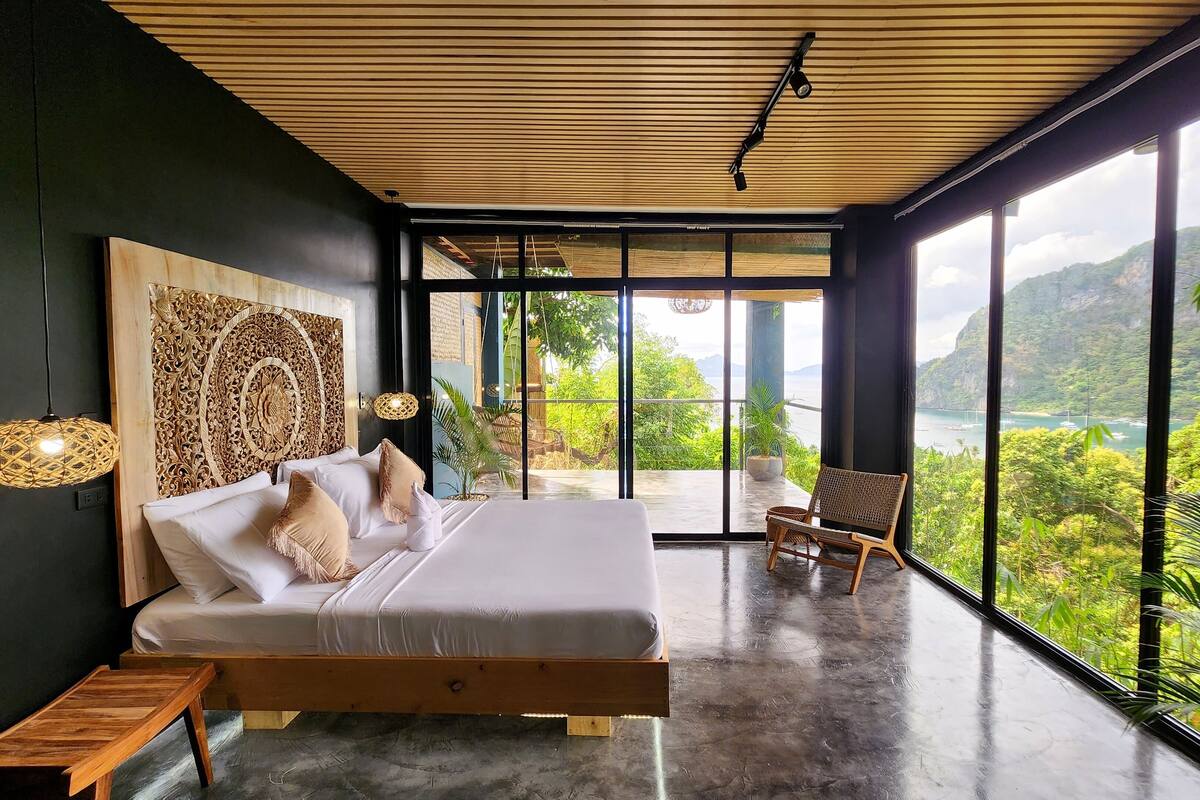 Ein Schlafzimmer mit einem großen Bett, einer hölzernen Bank und Blick auf eine gebirgige Landschaft durch große Fenster.