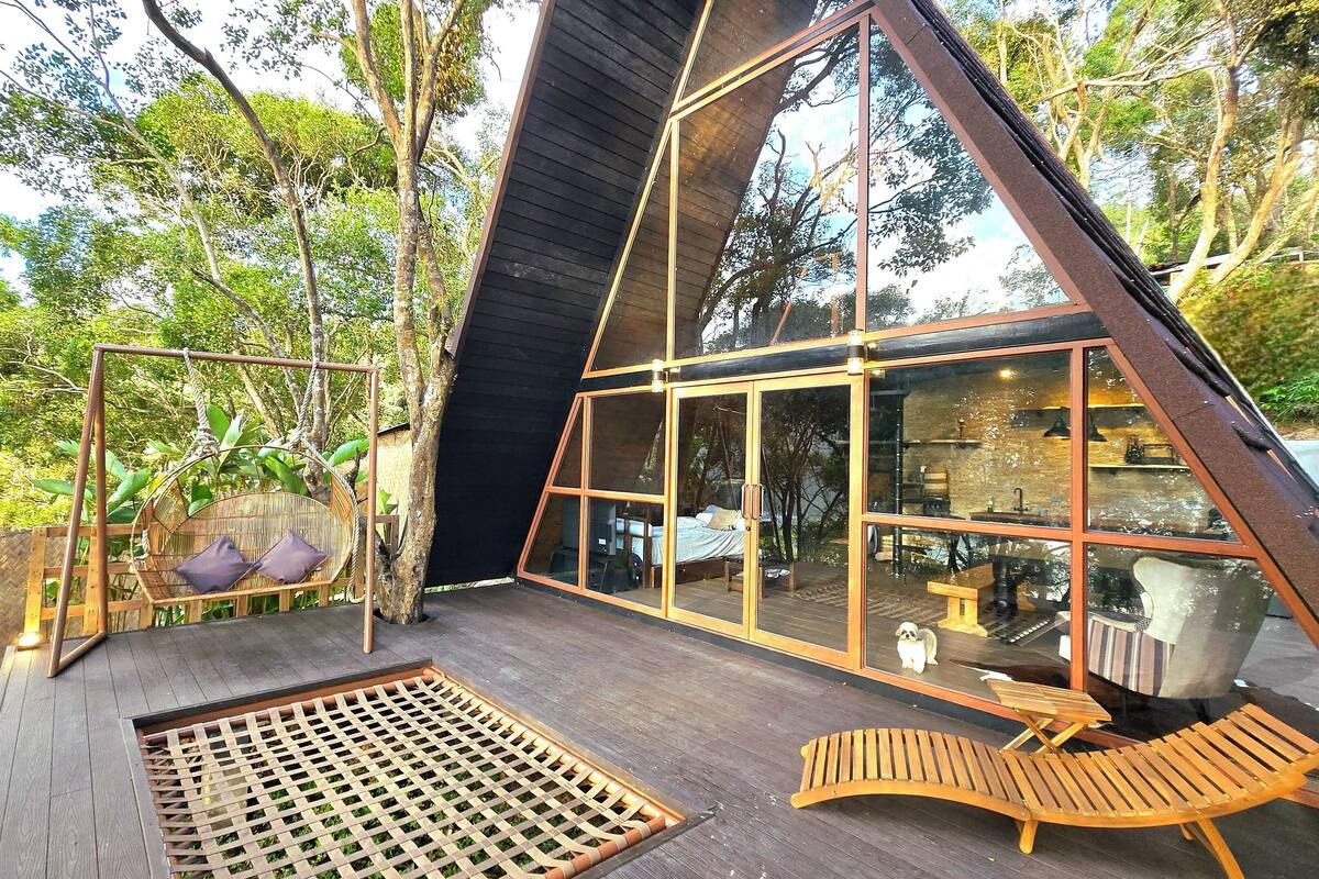 Eine moderne, dreieckige Holzkonstruktion mit großen Glasfenstern, einer Holzterrasse und einem Hängekorbstuhl.