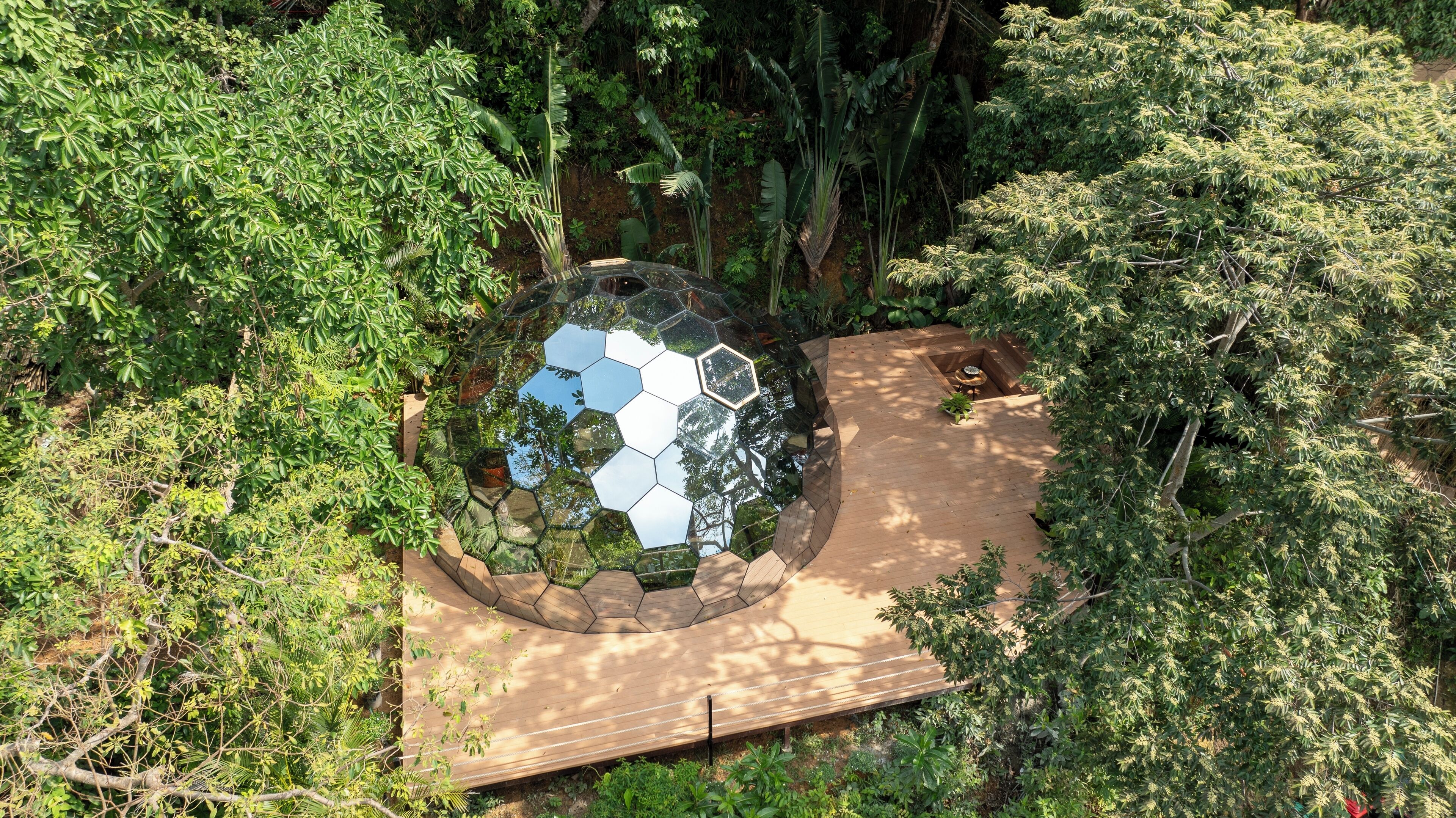 A hexagonal glass structure surrounded by a wooden deck, set in a lush garden with dense foliage.