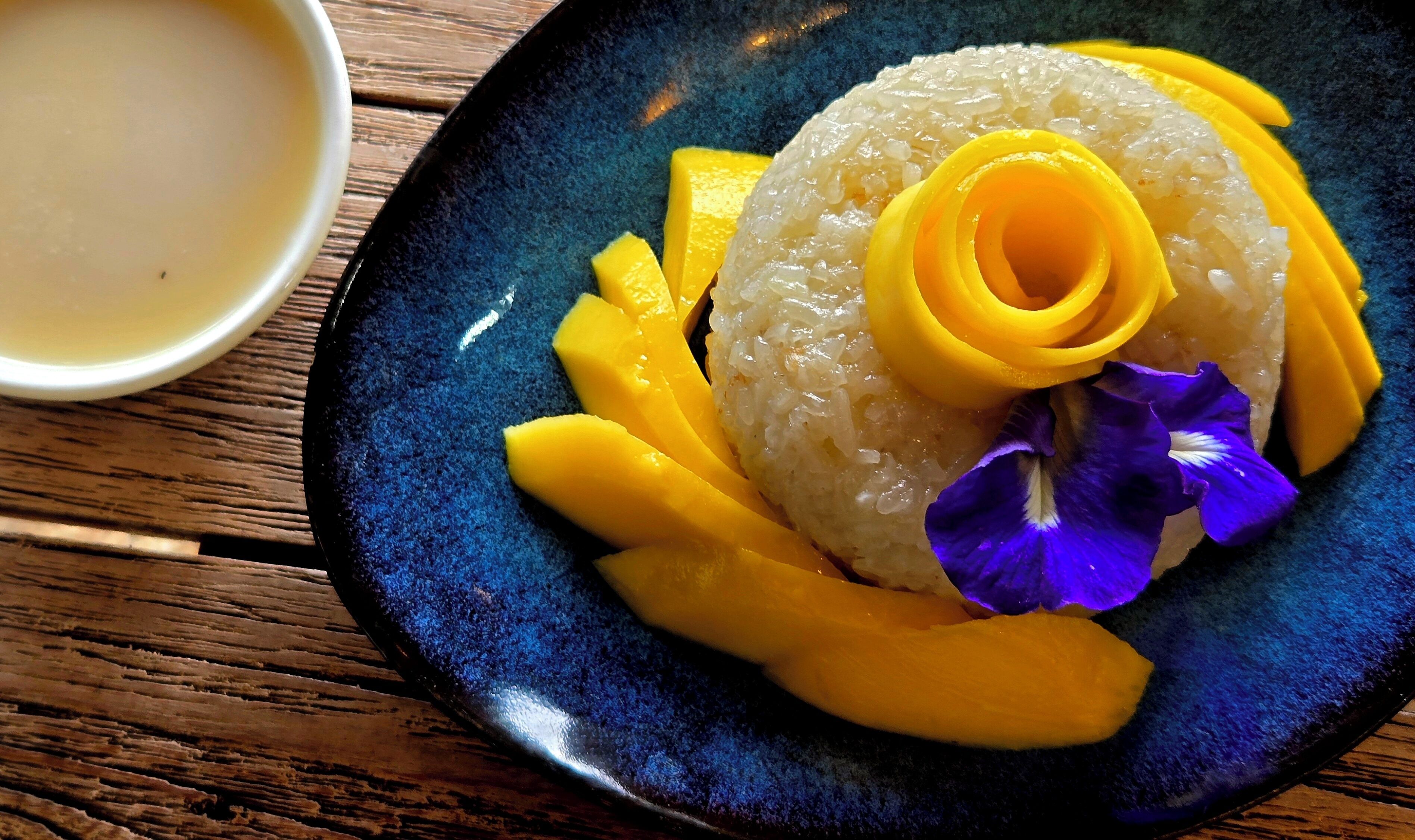 A plate of rice topped with mango slices and a purple flower, accompanied by a small bowl of soup and chopsticks on a wooden table.