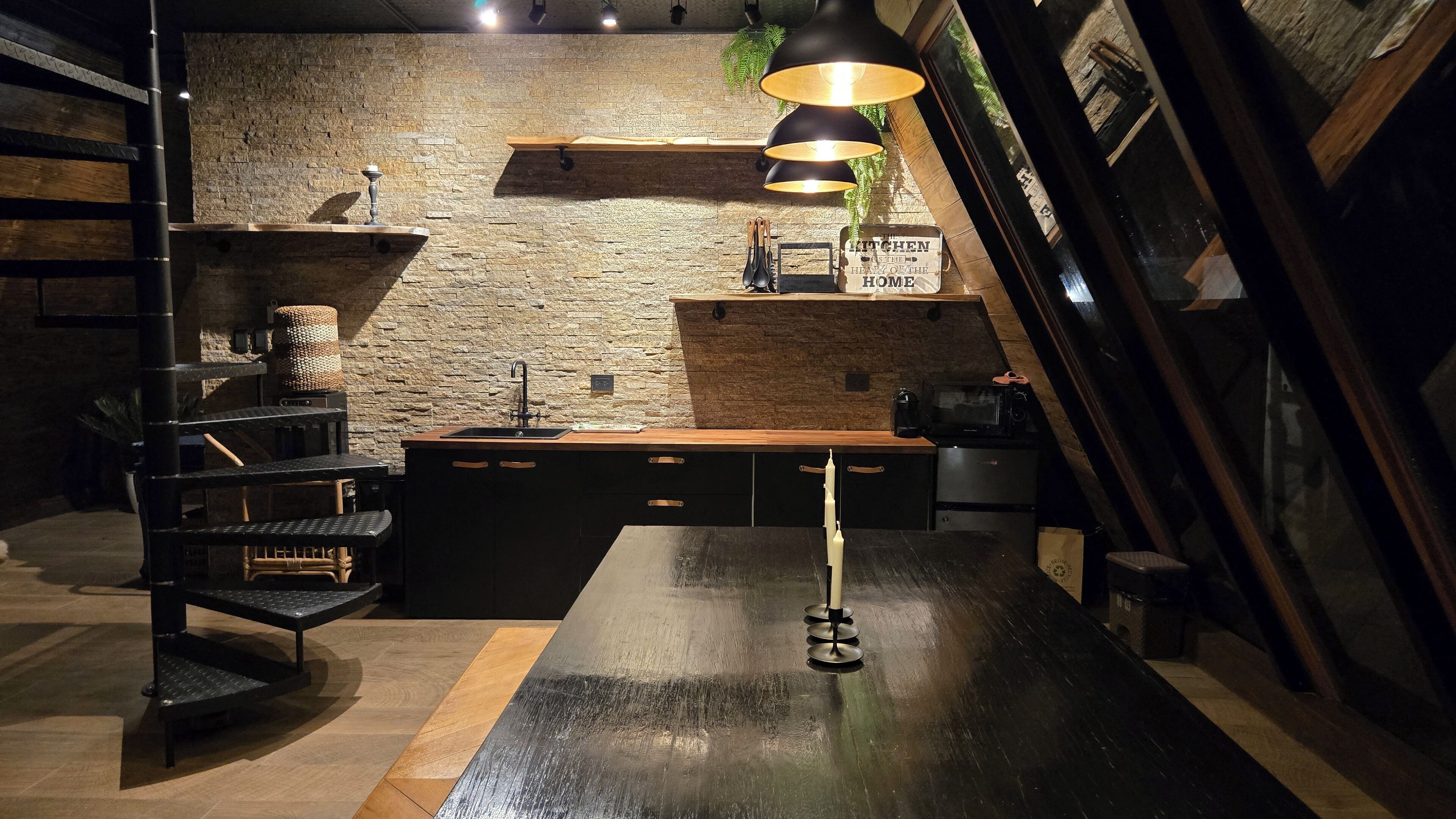 A modern kitchen with a stone wall, wooden shelves, a black countertop, and a hanging light fixture.