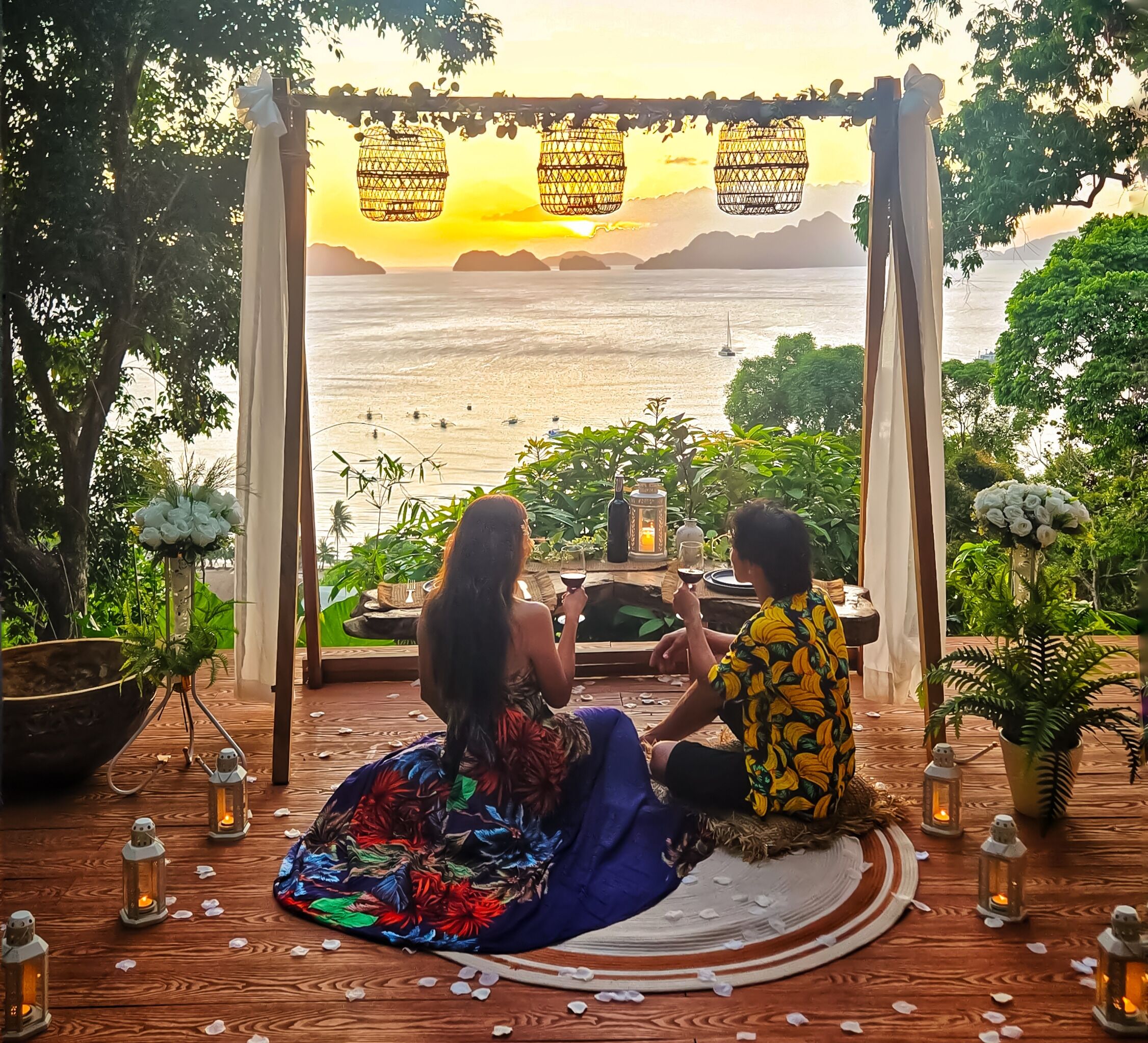 A couple sitting on a wooden deck overlooking a beach with a sunset view, surrounded by greenery and decorative lanterns.