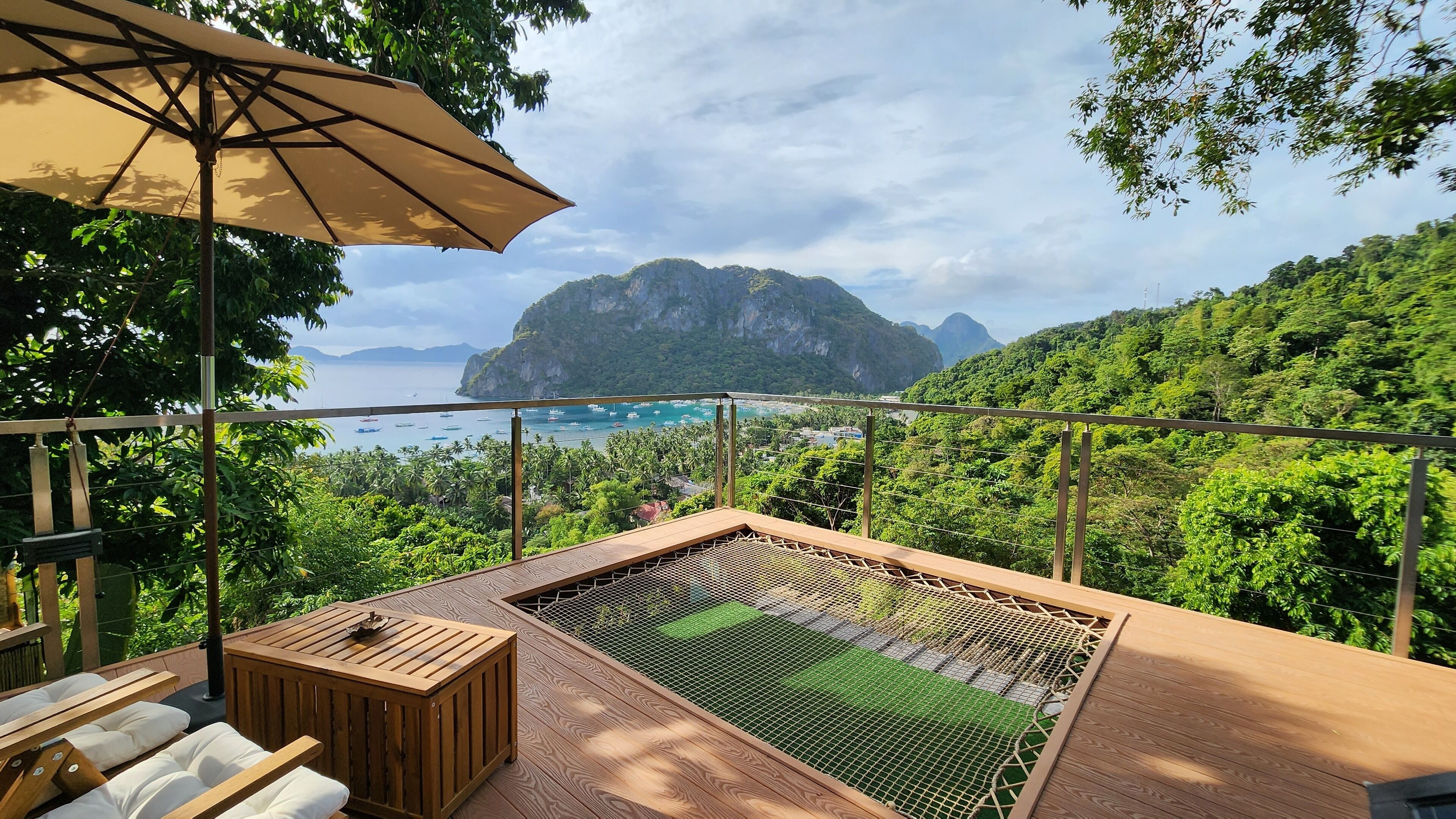 Una terraza de madera con zona de estar y jacuzzi, con vistas a un paisaje costero con barcos y abundante vegetación.