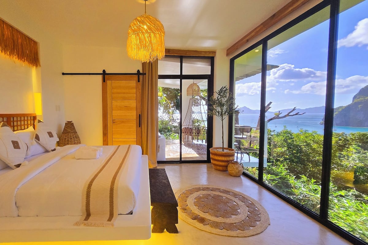 Ein Schlafzimmer mit einem großen Bett, einer hängenden Leuchte und Blick auf eine Küstenlandschaft durch eine Glastür.