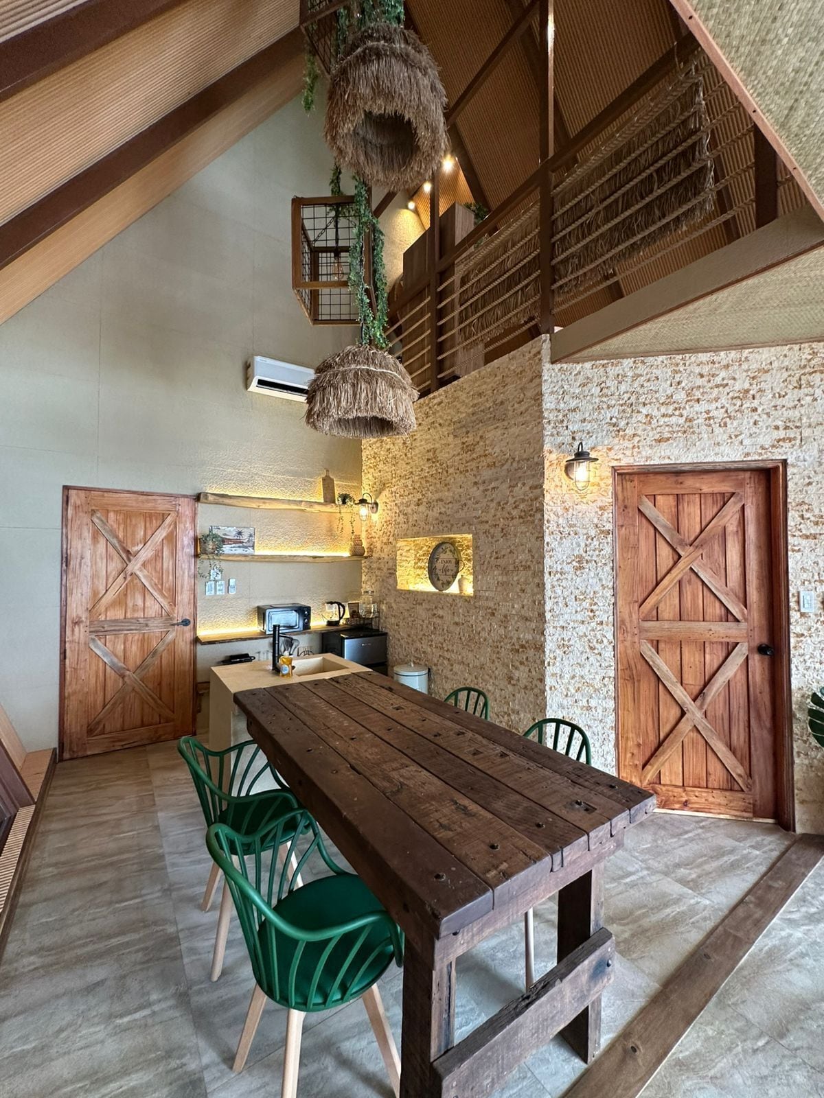 A rustic kitchen with a wooden table and chairs, a hanging light fixture, and a stone wall.
