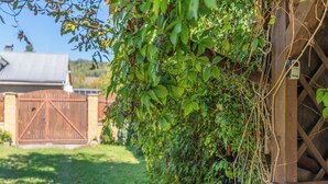 Une clôture en bois ornée de plantes grimpantes, un portail en métal et, au fond, un bâtiment doté d’un toit en pente.