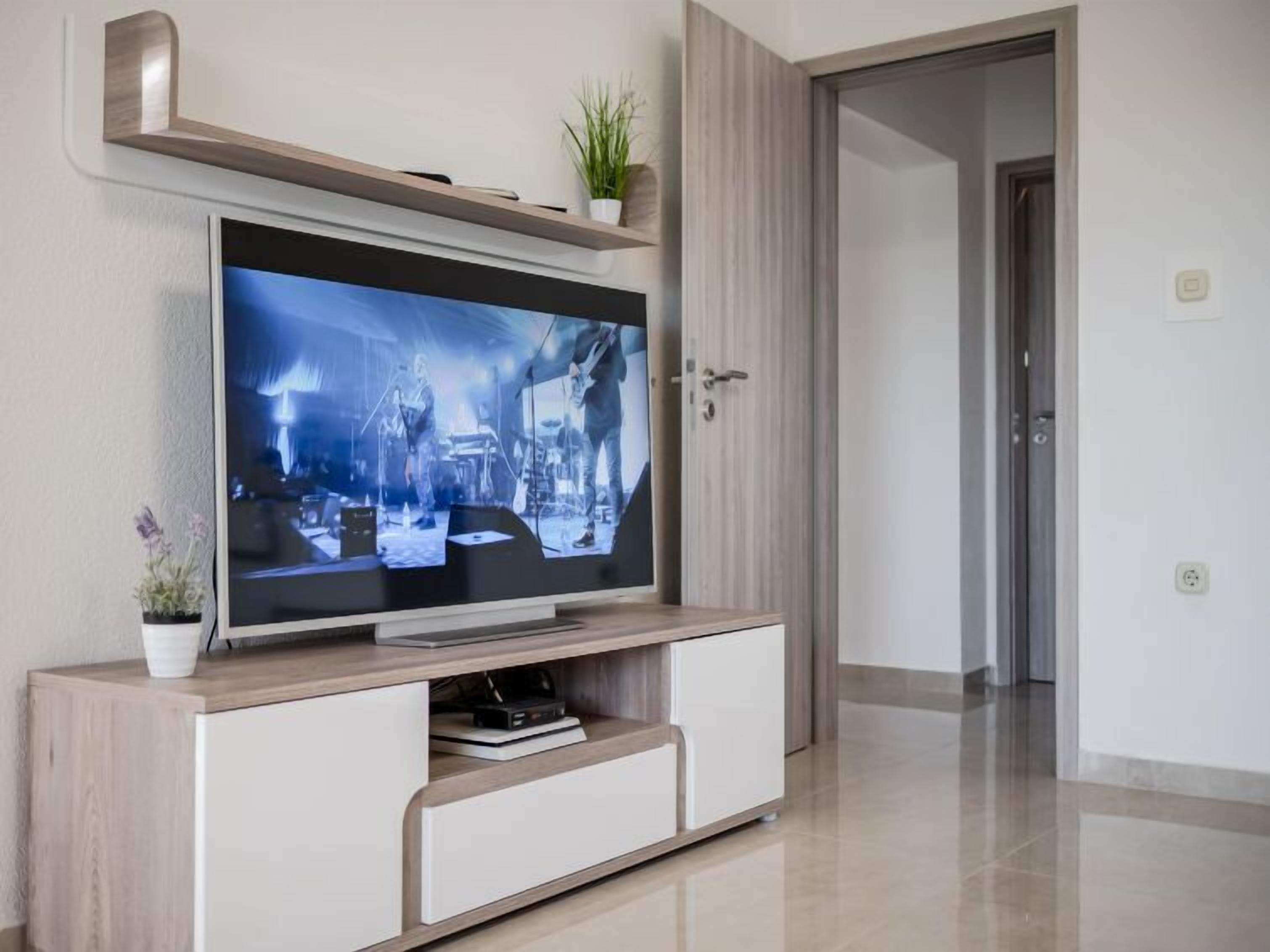 A modern living room with a flat-screen TV mounted on a wooden cabinet, a potted plant on the cabinet, and a door leading to another room.