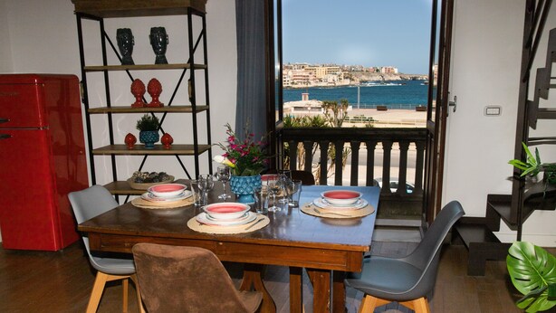 A dining area with a view of the sea, a red refrigerator, and a wooden table set for two.
