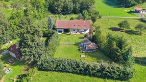 Une grande maison avec un toit recouvert de tuiles rouges, entourée de champs verdoyants et d’arbres.