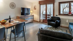 A modern living room with a dining area, a flat-screen TV, a sofa, and a window with curtains. - Condominio Giulia by Interhome (Canazei)