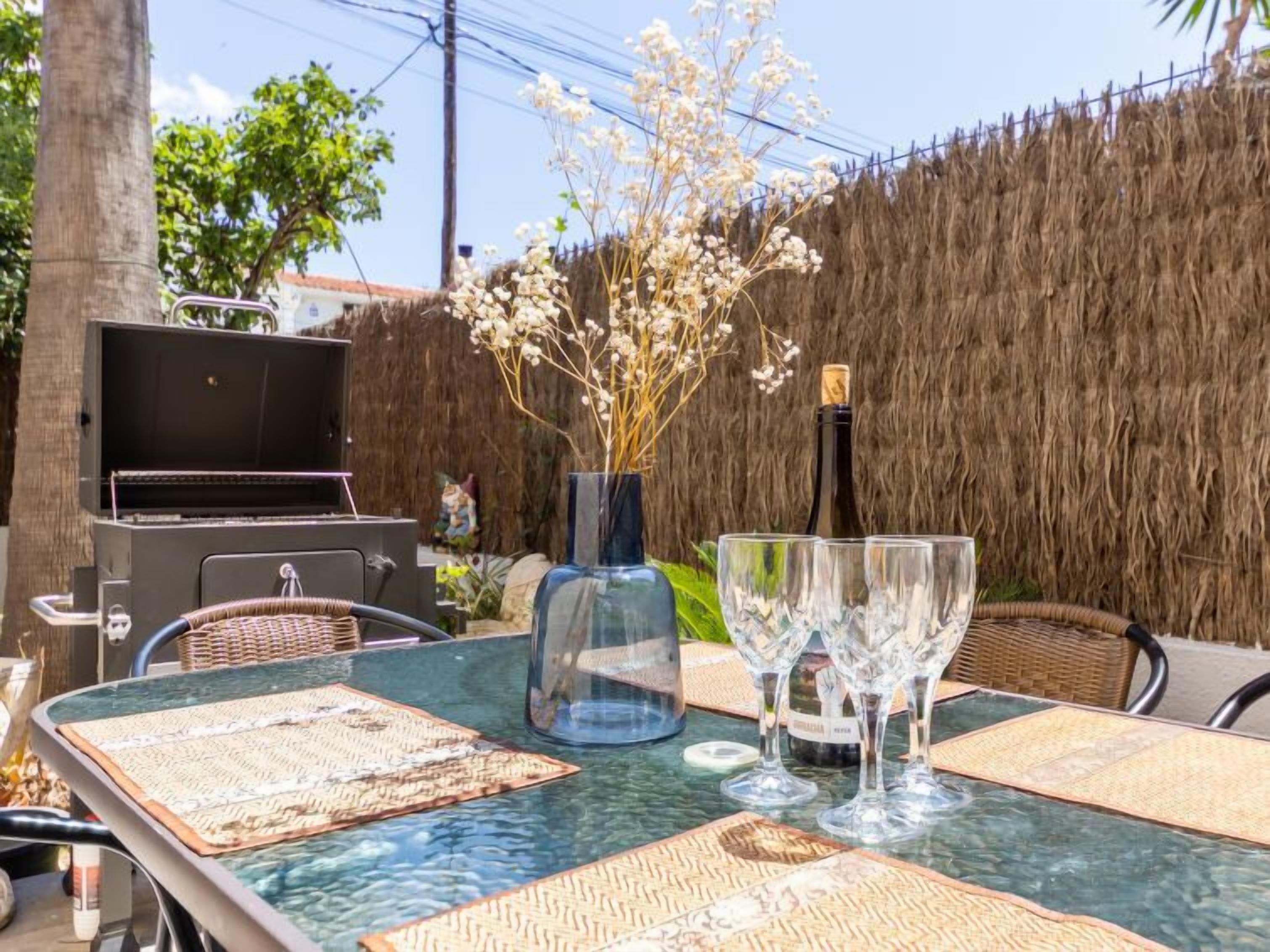 Un área de comedor al aire libre con una mesa de vidrio, sillas de mimbre, una parrilla y un florero decorativo con flores.