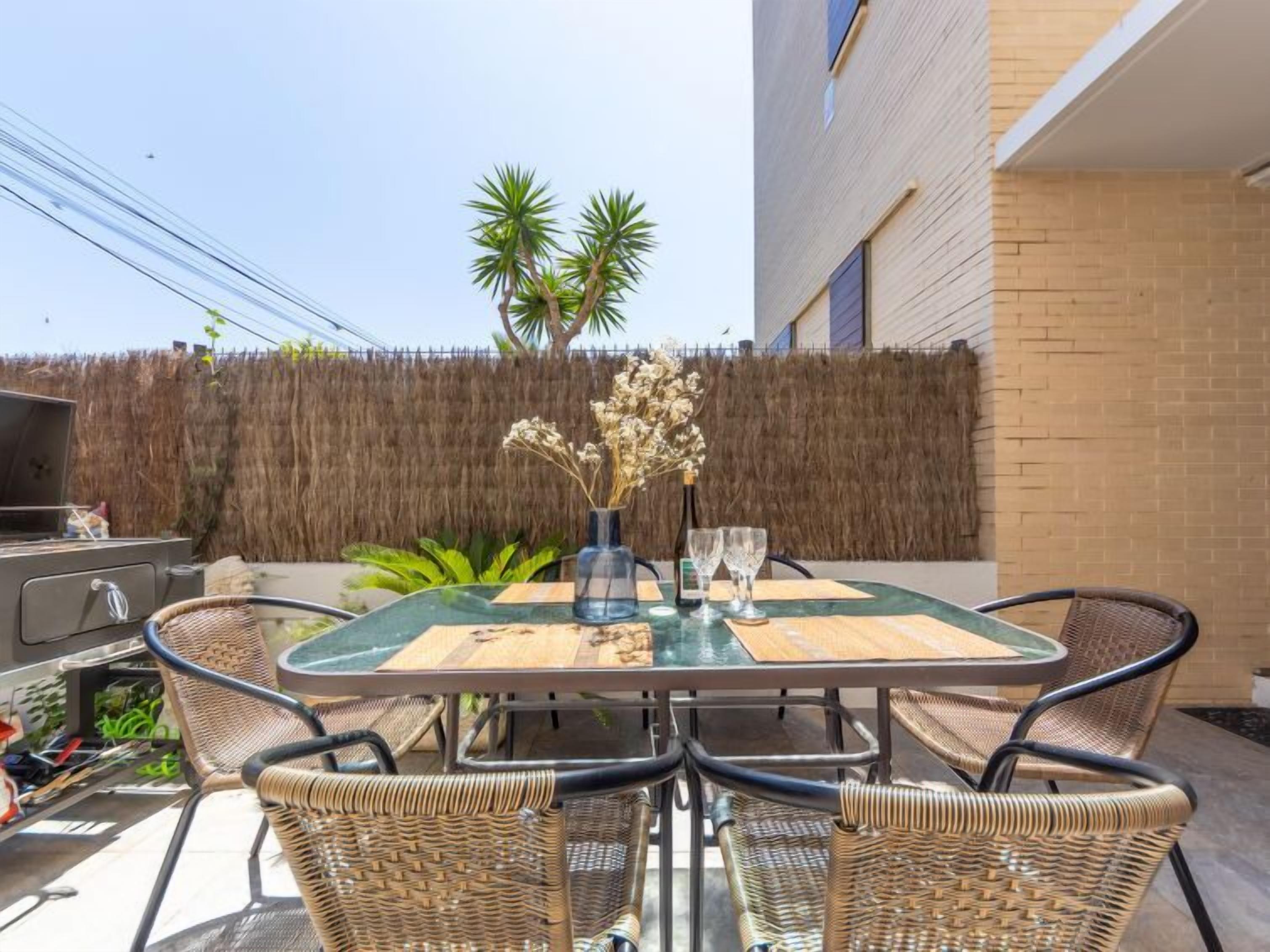 Un patio al aire libre con una mesa de vidrio, sillas de mimbre, una parrilla y un jarrón decorativo.