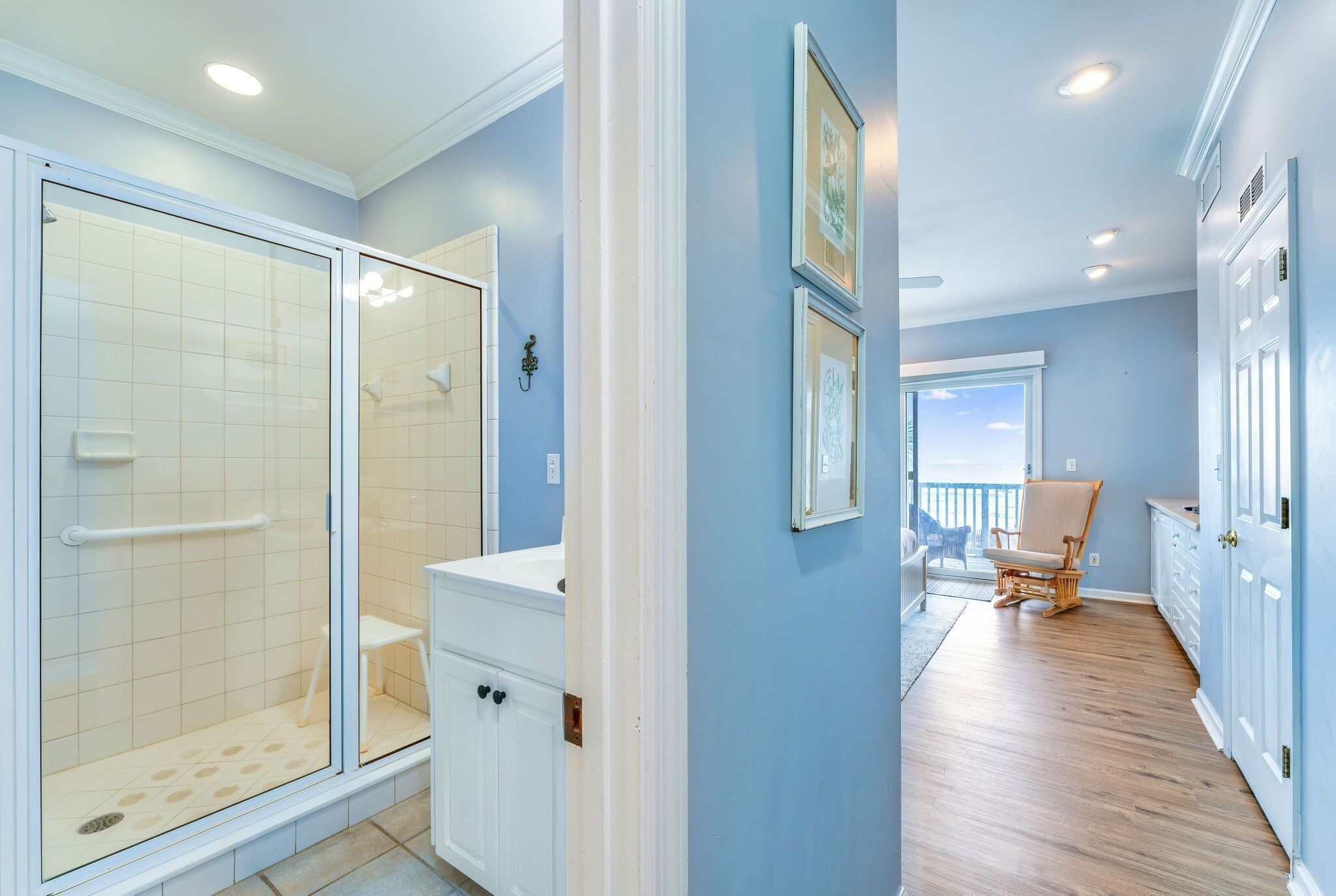 A bathroom with a glass shower enclosure, a white vanity, and a wooden chair in a room with blue walls and wooden flooring.