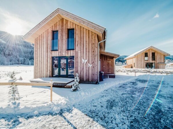 A wooden cabin with a large glass door and windows, set in a snowy landscape with mountains in the background.