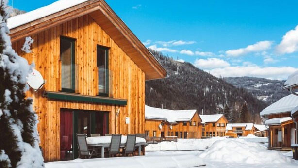 A wooden lodge with multiple rooms, a covered outdoor seating area, and a snowy mountain backdrop.