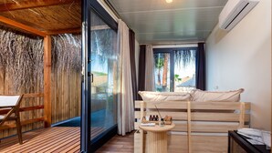 Una habitación con cama de madera, una mesita redonda con artículos de aseo, una silla y un amplio ventanal corredero que da a un área exterior.